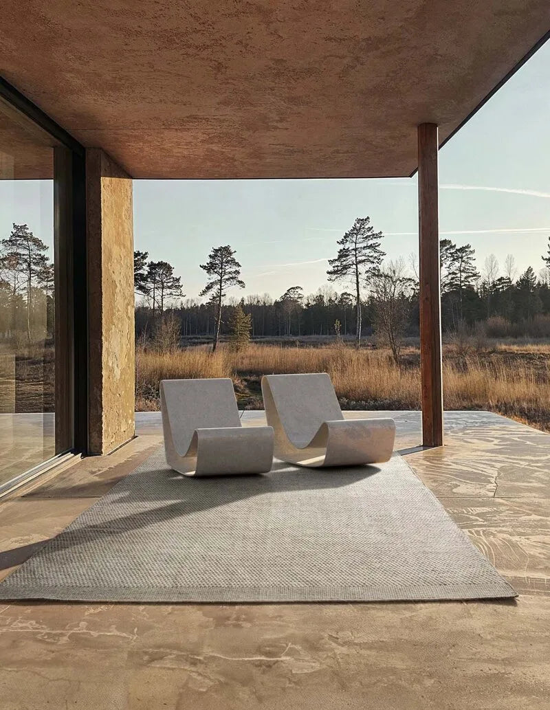 Heller, gewebter Outdoor-Teppich auf Steinterrasse mit zwei modernen weißen Loungesesseln vor weiter Naturlandschaft in ruhiger, minimalistischer Atmosphäre.