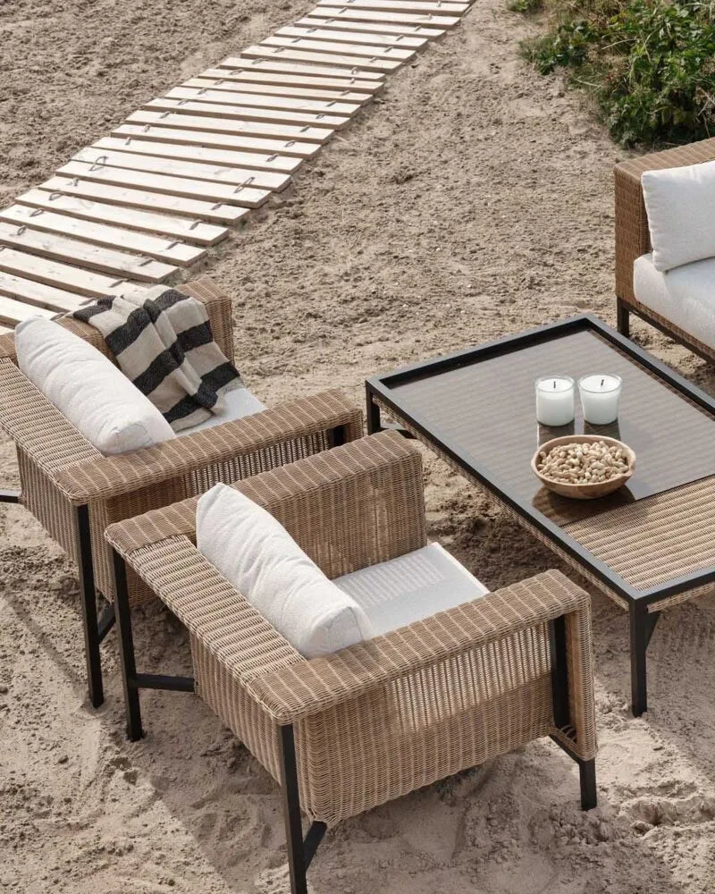 Zwei Outdoor-Sessel und ein Couchtisch aus Rattan und Metall mit hellen Kissen am sandigen Strand mit Holzsteg.