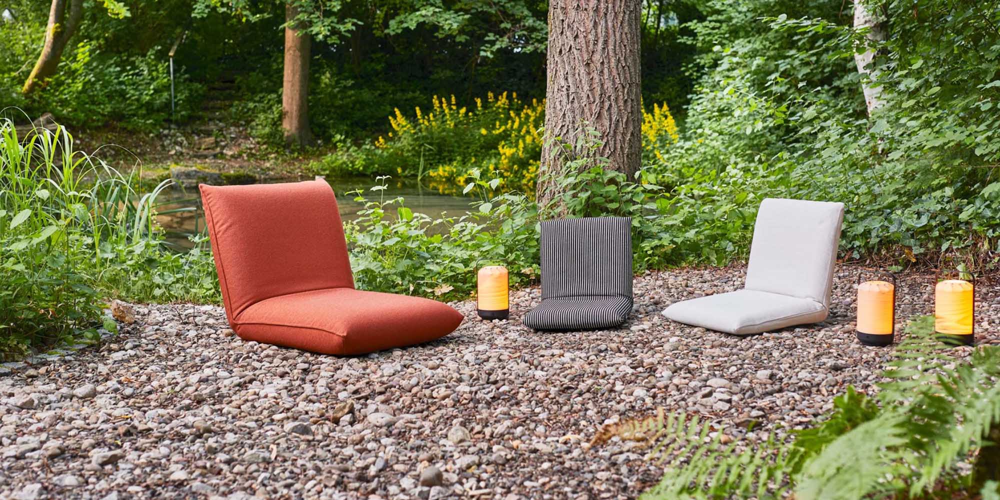 Drei gepolsterte Bodenstühle - ein roter, ein grauer und ein weißer - stehen auf einer Kiesfläche im Freien zwischen Bäumen und Grünpflanzen, und neben den Stühlen stehen zwei batteriebetriebene Laternen.