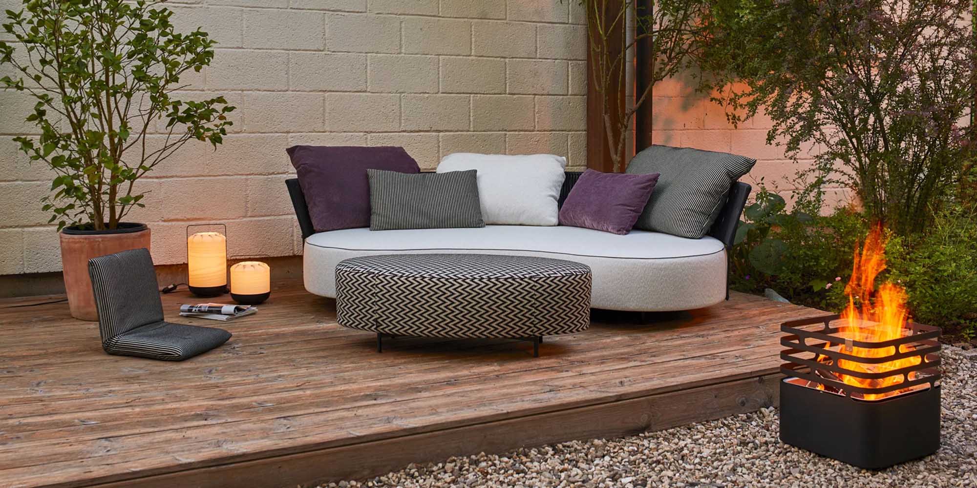 Eine gemütliche Außenterrasse mit einem geschwungenen weißen Sofa, verschiedenen Kissen, einer runden Ottomane, zwei Kissen auf der Holzterrasse, leuchtenden Laternen, Topfpflanzen und einer kleinen Feuerstelle mit hellen Flammen neben einer Steinmauer.