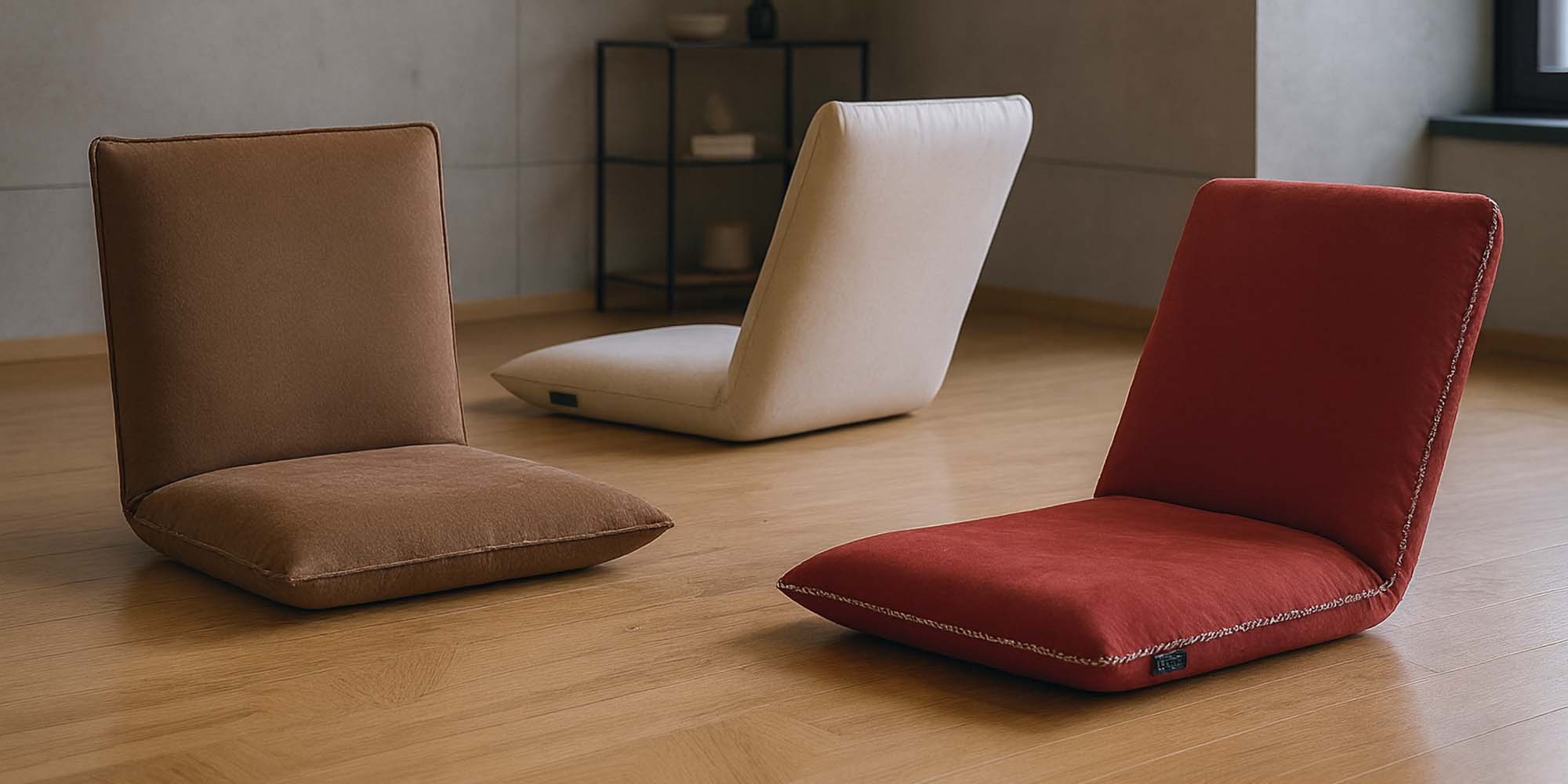 Drei Bodenstühle mit gepolsterter Sitzfläche und Rückenlehne - ein brauner, ein cremefarbener und ein roter - stehen in einem minimalistischen Raum mit Holzfußboden, einem kleinen Regal und einem Fenster.