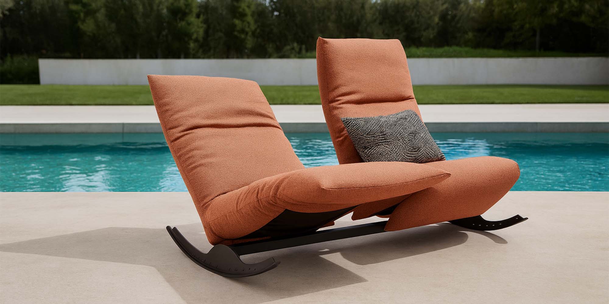 Zwei moderne orangefarbene Schaukelstühle mit weichen Polstern stehen nebeneinander auf einer Terrasse am Pool. Ein Stuhl hat ein graues, strukturiertes Kissen, und im Hintergrund sind ein klarer, blauer Swimmingpool und Grünpflanzen zu sehen.