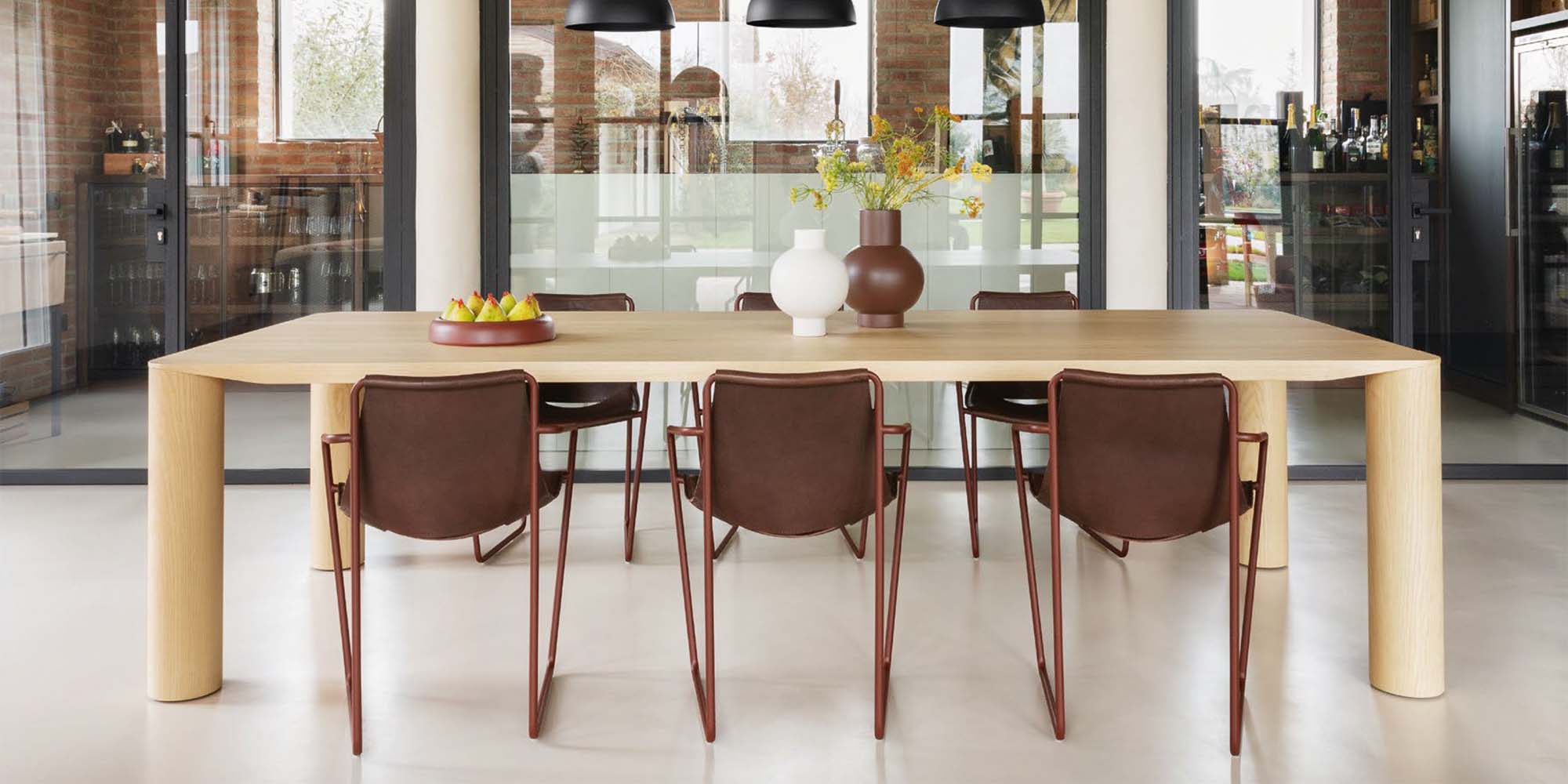 Ein modernes Esszimmer mit einem großen Holztisch, acht braunen Stühlen, einer Obstschale und zwei Vasen mit gelben Blumen. Im Hintergrund sind Glastüren und Backsteinwände zu sehen.