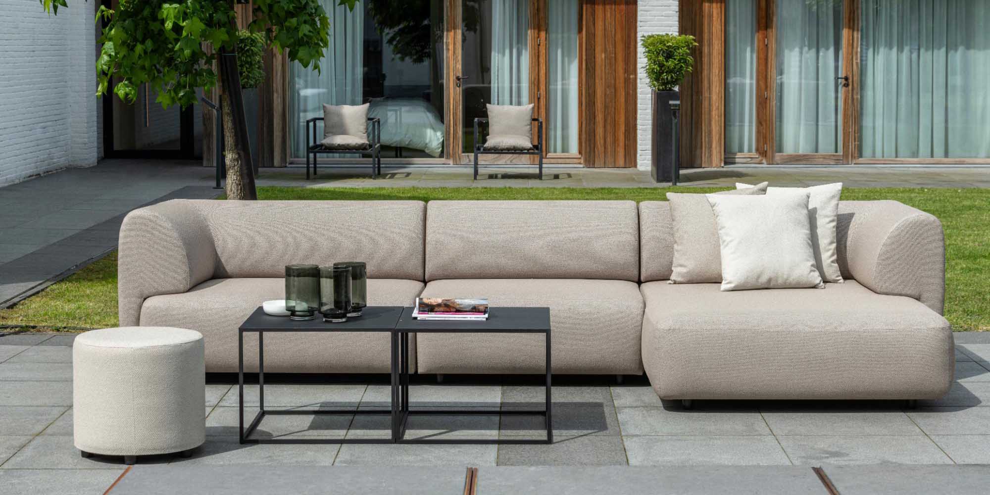 Eine moderne Außenterrasse mit einem beigen Sektionssofa und einer passenden Ottomane, schwarzen Tischen mit Gläsern und Zeitschriften und einer Kulisse aus Gras, Bäumen und Glastüren.