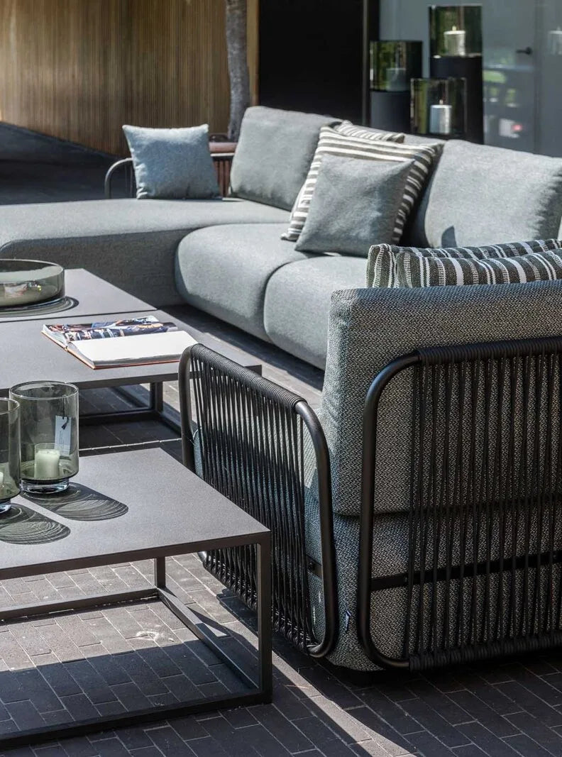 Modernes Outdoor-Loungesofa mit grauen Polstern und schwarzem Geflecht, kombiniert mit niedrigen Beistelltischen vor abendlicher Atmosphäre.