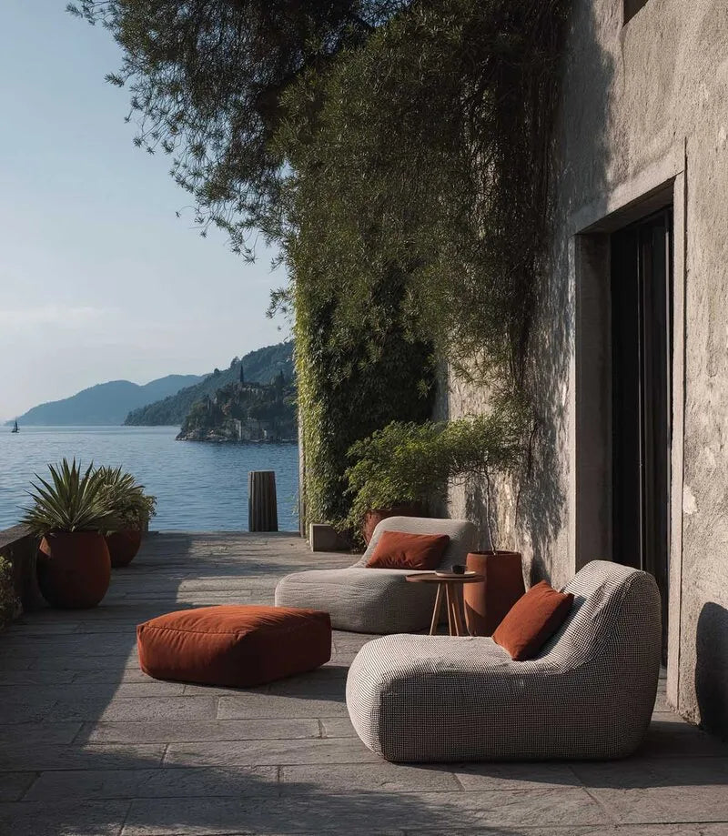 Zwei moderne Outdoor-Loungesessel in Beige mit orangefarbenem Hocker auf einer Steinterrasse vor mediterraner Küstenkulisse mit Felsen.