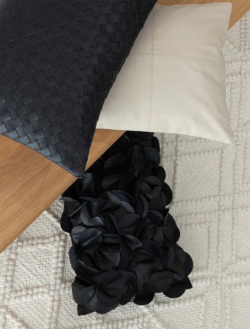 Drei moderne Kissen in Schwarz, Creme und mit floralem Volant-Bezug auf einem cremefarbenen Strukturteppich neben Holzelement.