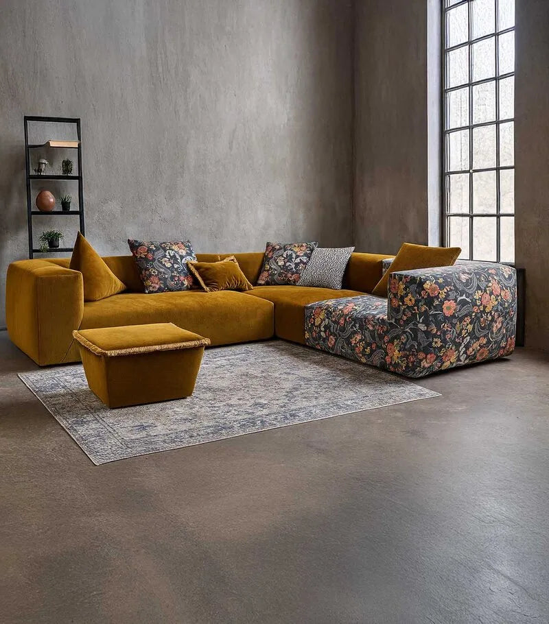 Modulares Ecksofa in Senfgelb mit floral gemustertem Bezug, kombiniert mit Kissen und Hocker in einem Betonraum.