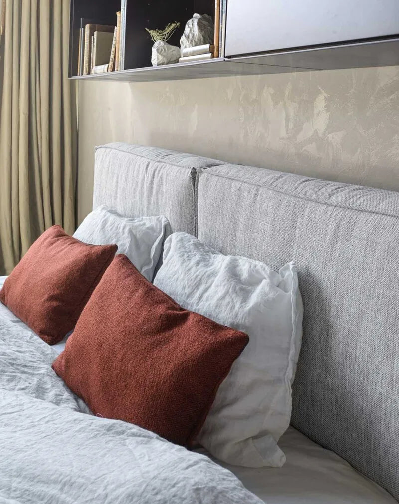 Polsterbett mit grauem Stoffbezug und abnehmbarer Husse, kombiniert mit weißer Leinenbettwäsche und rostroten Bouclé-Kissen in ruhigem Schlafzimmer.