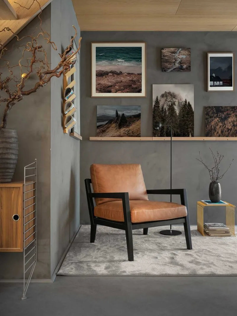 Cognac-farbener Ledersessel mit schwarzem Holzgestell vor graür Wand mit Bildergalerie, Stehlampe und hellem Teppich in moderner Atmosphäre.