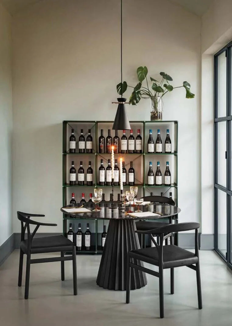 Eleganter runder Esstisch mit plissiertem Holzfuss, schwarzen Polsterstühlen und filigranem Weinregal vor heller Wand mit Tageslicht.