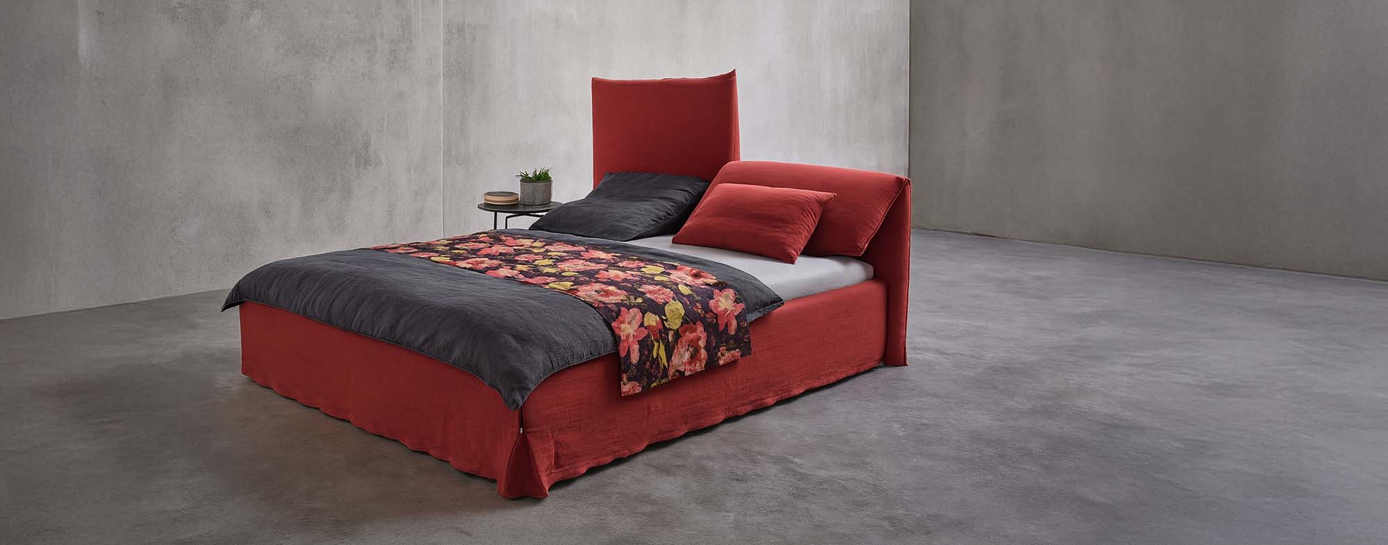 Ein modernes Bett mit einem rot gepolsterten Rahmen und Kopfteil, schwarzer Bettwäsche, einer geblümten Decke, roten und schwarzen Kissen und einem kleinen Beistelltisch mit einer Pflanze in einem minimalistischen Betonzimmer.