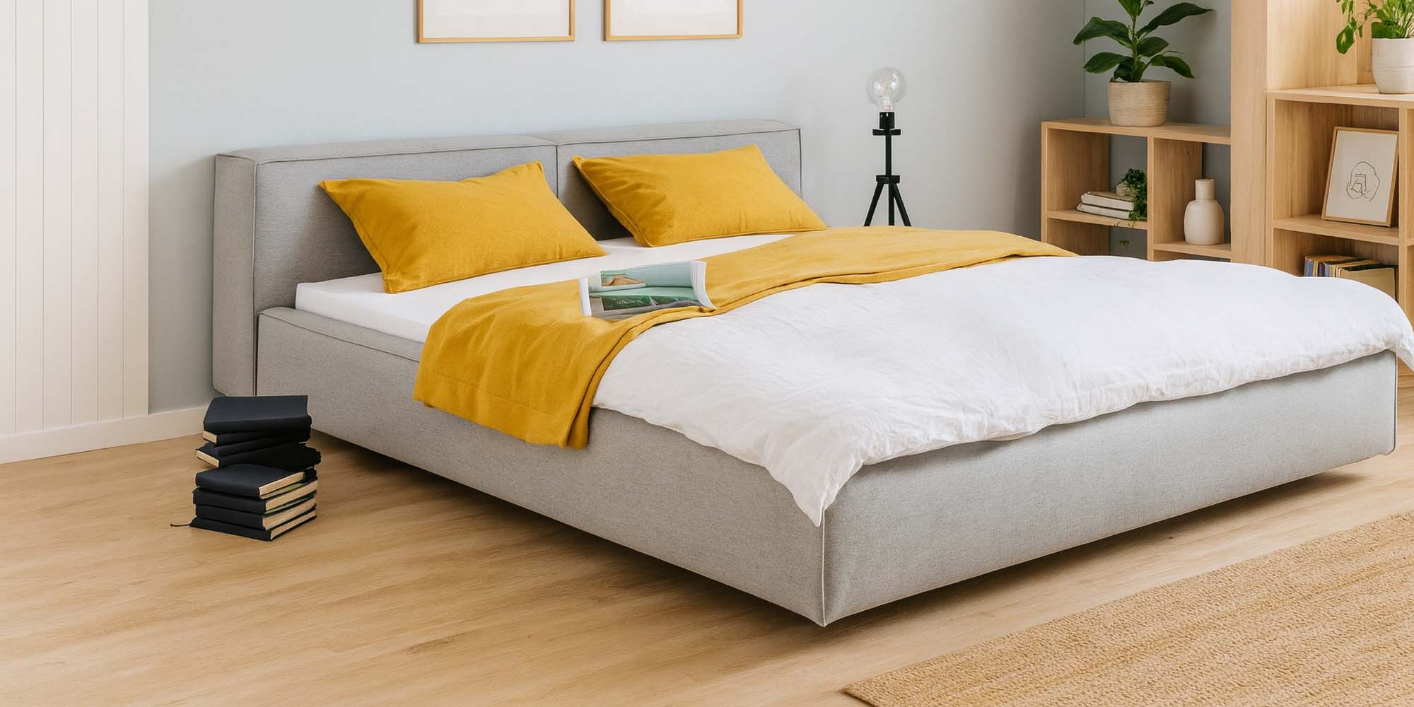 Ein modernes Schlafzimmer mit einem hellgrauen Polsterbett, weißer Bettwäsche und senfgelben Kissen und Überwürfen. Ein aufgeschlagenes Buch liegt auf dem Bett. Stapel von Büchern liegen auf dem Boden. Holzregale und Zimmerpflanzen schmücken den Raum.
