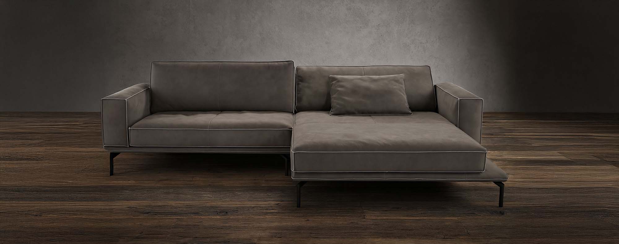 Ein modernes, dunkelgraues Sektionssofa mit Chaiselongue steht auf einem Holzboden vor einer strukturierten grauen Wand. Das Sofa hat klare Linien, Metallbeine und enthält ein passendes rechteckiges Kissen.