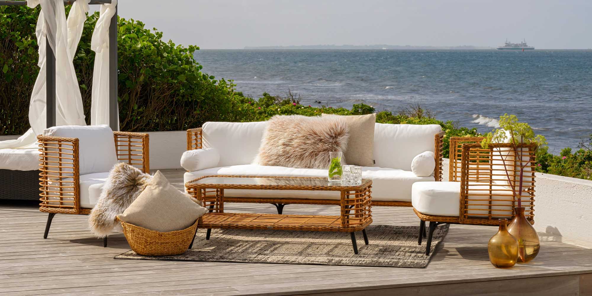 Terrasse im Freien mit Korbmöbeln, weißen Kissen, einem Teppich, dekorativen Kissen und Glasvasen. Die Einrichtung blickt auf den Ozean, im Hintergrund sind Grünpflanzen und ein Schiff zu sehen.