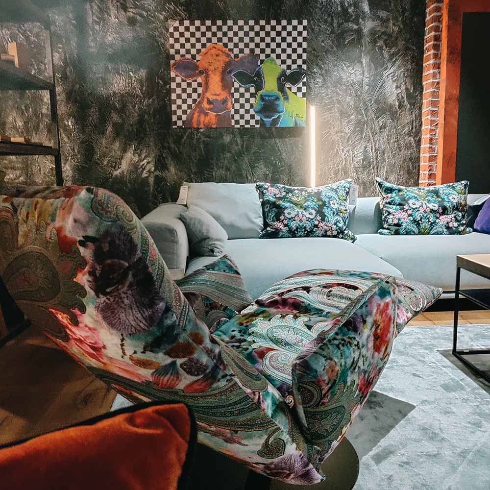 Ein modernes Wohnzimmer mit einem gemusterten Sessel, einem grauen Sofa mit Blumenkissen, einem Teppich und einem Wandbild mit bunten Kühen auf einem karierten Hintergrund. Die Wände haben eine strukturierte dunkle Oberfläche.