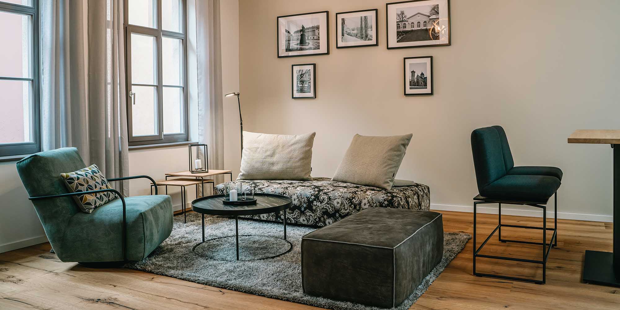 Modernes Wohnzimmer mit gemustertem Sofa, grünem Sessel, grauer Ottomane, kleinen runden Tischen, neutralem Teppich und gerahmten Schwarz-Weiß-Fotos an cremefarbenen Wänden, mit großen Fenstern, die natürliches Licht hereinlassen.